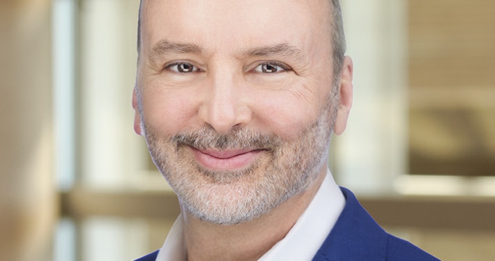Headshot of a man in a blue suit coat and white shirt in front of a bank of windows.