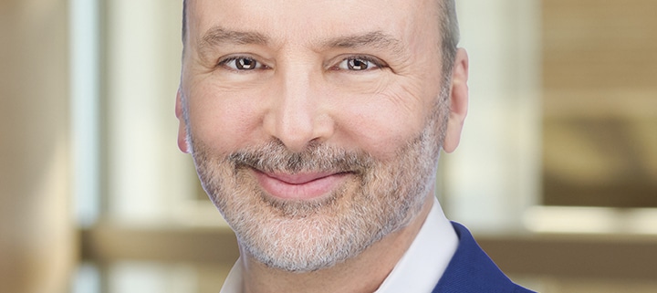 Headshot of a man in a blue suit coat and white shirt in front of a bank of windows.