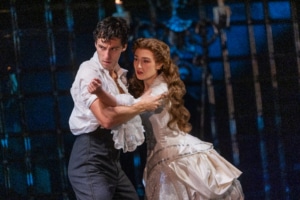 A dancing couple in period costumes moves together in a tense, emotional embrace on a dimly lit stage