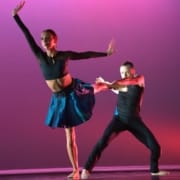Two dancers in front of a deep pink background. One with arms extended, standing on one leg while the other pulls the first dancers other leg backward.