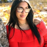 Person with long dark hair sitting against a tree in a park with autumn foliage, wearing a bright red off‑the‑shoulder sweater and layered necklaces.