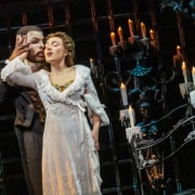 Two performers stand close in a candlelit Gothic setting, one in a flowing white gown and the other in dark formal attire, creating an intimate moment on stage.