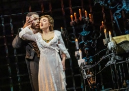 Two performers stand close in a candlelit Gothic setting, one in a flowing white gown and the other in dark formal attire, creating an intimate moment on stage.