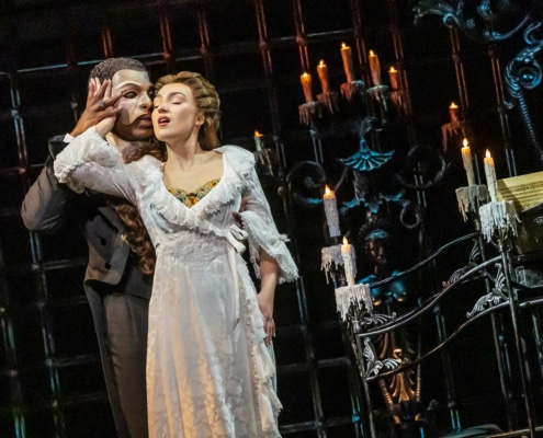 Two performers stand close in a candlelit Gothic setting, one in a flowing white gown and the other in dark formal attire, creating an intimate moment on stage.