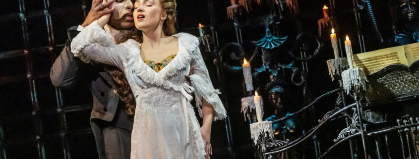 Two performers stand close in a candlelit Gothic setting, one in a flowing white gown and the other in dark formal attire, creating an intimate moment on stage.