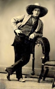 A black and white photograph of Jack Langrishe from the 1800s, shown standing against a chair with a large brimmed hat and western attire.
