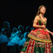 A soprano in an elaborate red and green gown stands in the foreground while a line of dancers in pale costumes echoes her pose behind her.”