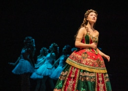 A soprano in an elaborate red and green gown stands in the foreground while a line of dancers in pale costumes echoes her pose behind her.”