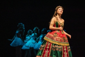 A soprano in an elaborate red and green gown stands in the foreground while a line of dancers in pale costumes echoes her pose behind her.”