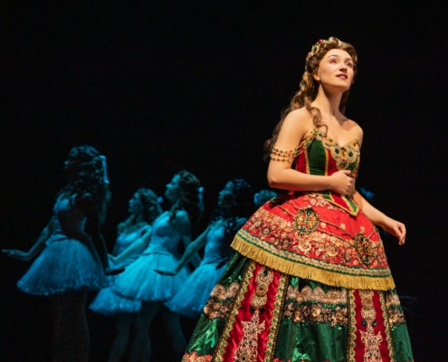 A soprano in an elaborate red and green gown stands in the foreground while a line of dancers in pale costumes echoes her pose behind her.”