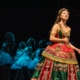 A soprano in an elaborate red and green gown stands in the foreground while a line of dancers in pale costumes echoes her pose behind her.”