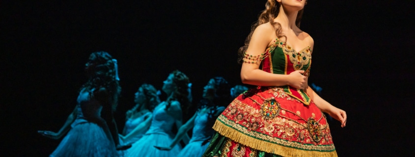 A soprano in an elaborate red and green gown stands in the foreground while a line of dancers in pale costumes echoes her pose behind her.”