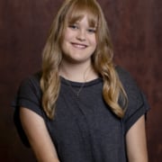 Person with wavy blonde hair wearing a dark gray top and gold necklace, seated in front of a dark studio backdrop.