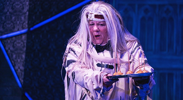 Actress dressed in a white straight jacket with long, white wig holding out a silver tray with appetizers