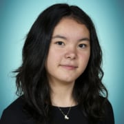Person with wavy dark hair wearing a black top and a small pendant necklace, photographed against a teal studio backdrop.