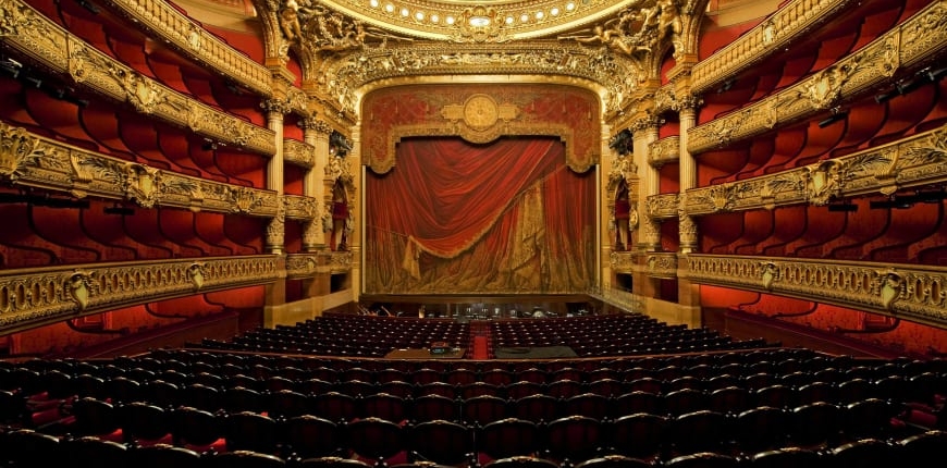 A color photo of an opulent opera house designed in tones of red and gold with side boxes, a red curtain, and ornate ceiling.
