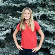 Person with long blonde hair standing outdoors in front of a large evergreen tree, wearing a red collared shirt and a dark plaid skirt with hands on hips.