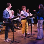 A group of actors onstage with music stands rehearsing a reading, as a director stands in front giving direction.