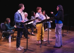 A group of actors onstage with music stands rehearsing a reading, as a director stands in front giving direction.