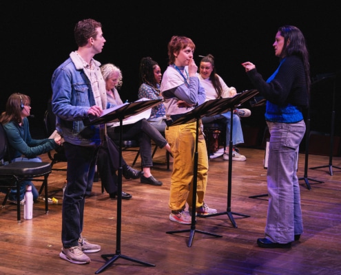 A group of actors onstage with music stands rehearsing a reading, as a director stands in front giving direction.