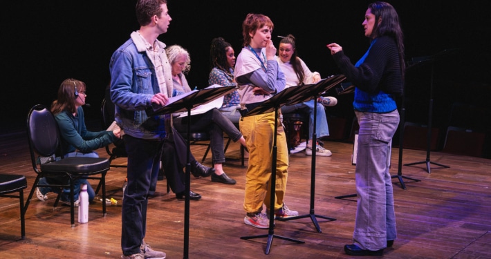A group of actors onstage with music stands rehearsing a reading, as a director stands in front giving direction.