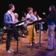 A group of actors onstage with music stands rehearsing a reading, as a director stands in front giving direction.