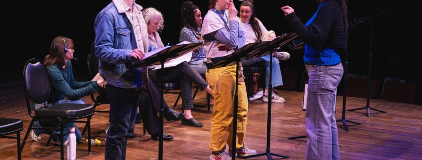 A group of actors onstage with music stands rehearsing a reading, as a director stands in front giving direction.