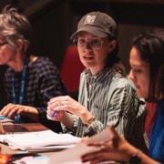 Three people seated at a table in a theater space, reviewing scripts and taking notes during a rehearsal meeting.