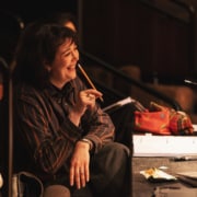 Person sitting at a rehearsal table in a dimly lit theater, holding a pencil and observing the stage.