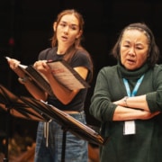 Two people standing onstage with music stands, one holding a script and the other standing with arms crossed during rehearsal.