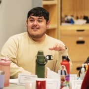 Person speaking during a table read, gesturing while surrounded by scripts, water bottles, and rehearsal materials.
