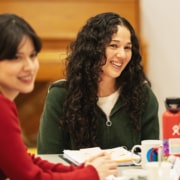 Two people sitting at a table in a rehearsal room, discussing notes with open scripts and coffee mugs in front of them.