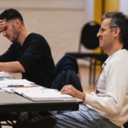 Two people seated at a long table in a rehearsal studio, reading through materials and taking notes.