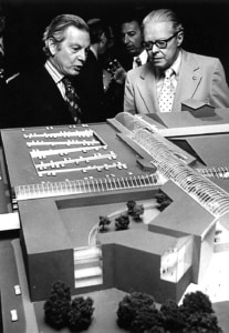 Black‑and‑white photo of two people viewing an architectural scale model featuring multiple connected buildings, walkways, and landscaped areas.