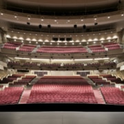 Wide view of a large, modern theater auditorium with multiple balconies and rows of red seats, seen from the stage looking out toward the house.
