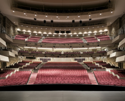 Wide view of a large, modern theater auditorium with multiple balconies and rows of red seats, seen from the stage looking out toward the house.