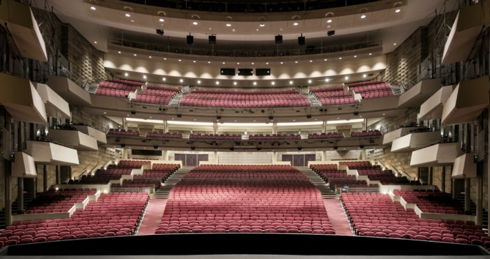 Wide view of a large, modern theater auditorium with multiple balconies and rows of red seats, seen from the stage looking out toward the house.