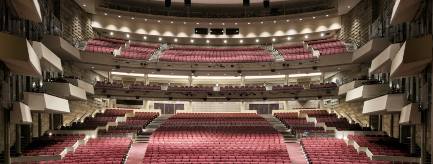 Wide view of a large, modern theater auditorium with multiple balconies and rows of red seats, seen from the stage looking out toward the house.