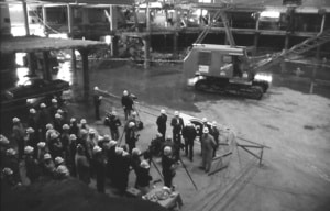 Historical black‑and‑white image of a construction site where a group of workers in hard hats gather near equipment inside a large partially built structure.