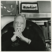 Black‑and‑white photo of a person seated in a leather chair with hands clasped, surrounded by bookshelves displaying art books, photographs, and theater-related materials.