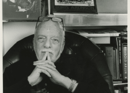 Black‑and‑white photo of a person seated in a leather chair with hands clasped, surrounded by bookshelves displaying art books, photographs, and theater-related materials.
