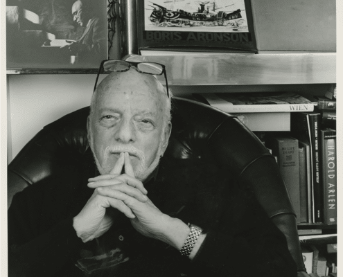 Black‑and‑white photo of a person seated in a leather chair with hands clasped, surrounded by bookshelves displaying art books, photographs, and theater-related materials.