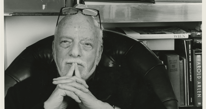 Black‑and‑white photo of a person seated in a leather chair with hands clasped, surrounded by bookshelves displaying art books, photographs, and theater-related materials.