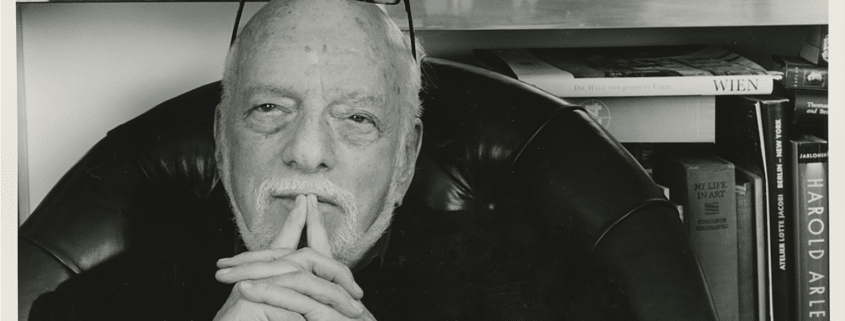 Black‑and‑white photo of a person seated in a leather chair with hands clasped, surrounded by bookshelves displaying art books, photographs, and theater-related materials.