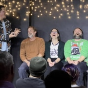 Performer gesturing animatedly while three seated participants take part in an improv or comedy scene before a live audience, with string lights hanging behind them.