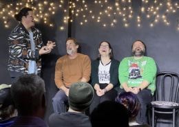 Performer gesturing animatedly while three seated participants take part in an improv or comedy scene before a live audience, with string lights hanging behind them.
