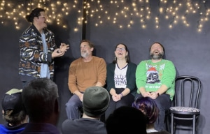Performer gesturing animatedly while three seated participants take part in an improv or comedy scene before a live audience, with string lights hanging behind them.