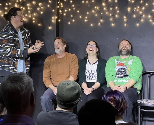 Performer gesturing animatedly while three seated participants take part in an improv or comedy scene before a live audience, with string lights hanging behind them.