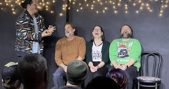 Performer gesturing animatedly while three seated participants take part in an improv or comedy scene before a live audience, with string lights hanging behind them.