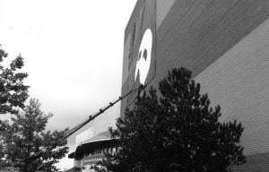 Black-and-white photo of a large banner featuring the Phantom of the Opera mask being installed on the side of a tall brick building using a crane.